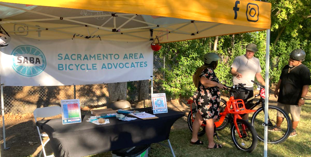 Under a canopy, 3 people look at an e-bike. There is a table with literature and a banner "Sacramento Area Bicycle Advocates."