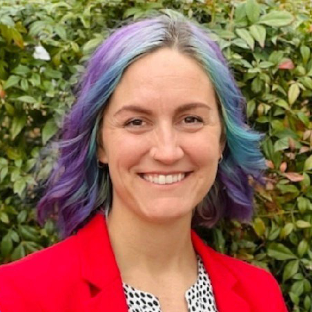 A white woman with blue & purple dyed hair and a red jacket smiles.