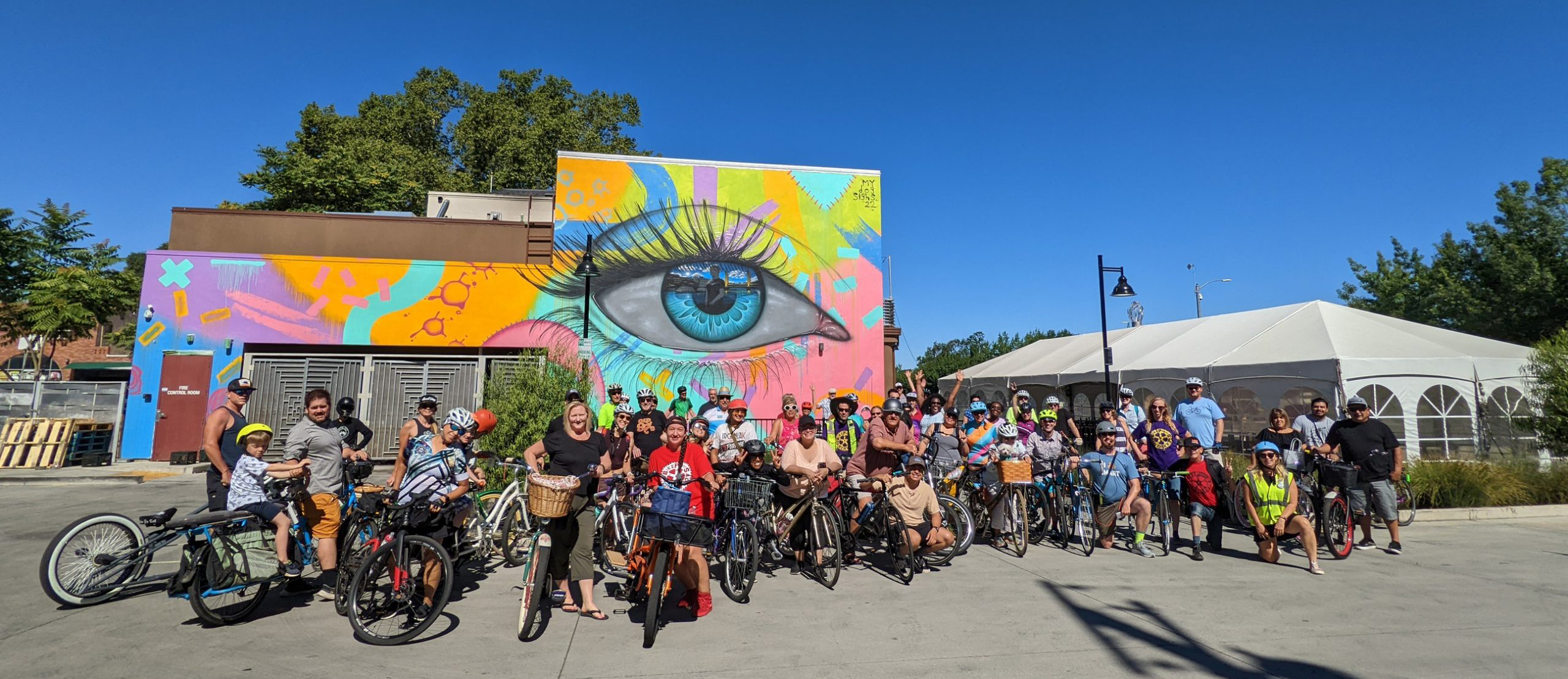 A large group of cyclists, including adults & children, pose in front of a mural of a large eye.