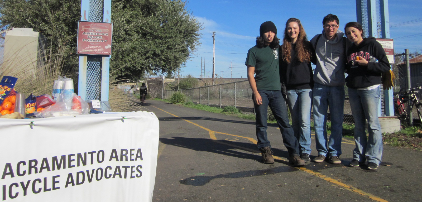 Four young people - two men and two women - stand on the American River Parkway next to a table that says "Sacramento Area Bicycle Advocates."