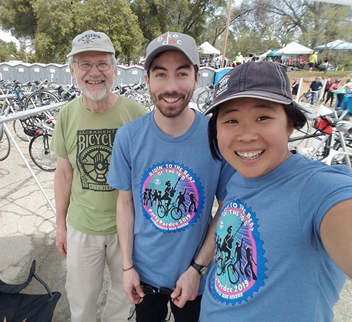 Three people - a young Asian woman, a young white man, and an older white man with bikes parked behind them.