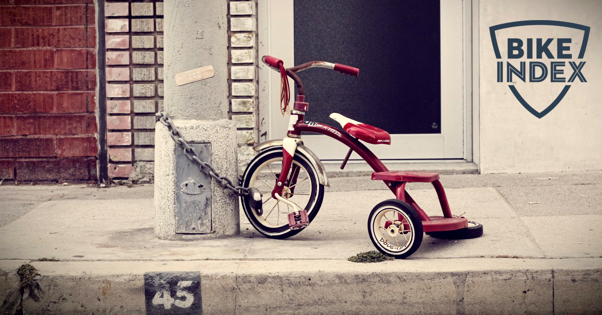 Outside a building, a red trike is chained to a concrete post. With Bike Index logo.