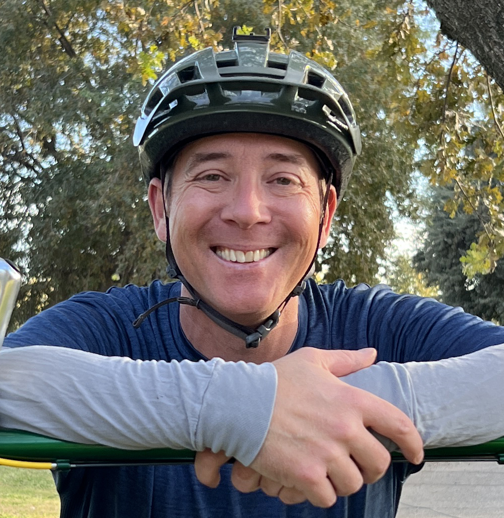 A man wearing a bike helmet leans on his bike.