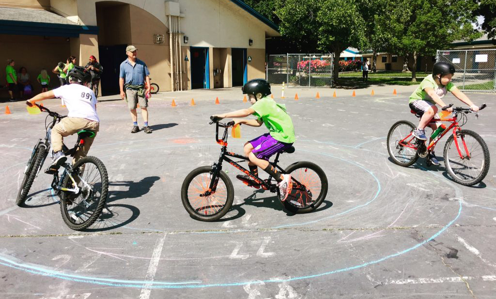 Kids on bikes ride around a chalked circle in a parking lot while an adult looks on.