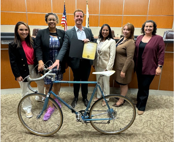 Six people pose in city council chambers, one man is holding a bike in front of him.