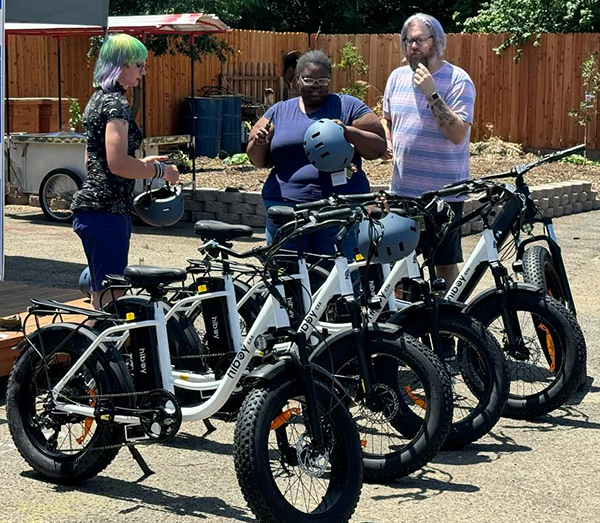 Three people holding bike helmets stand next to a row of e-bikes.