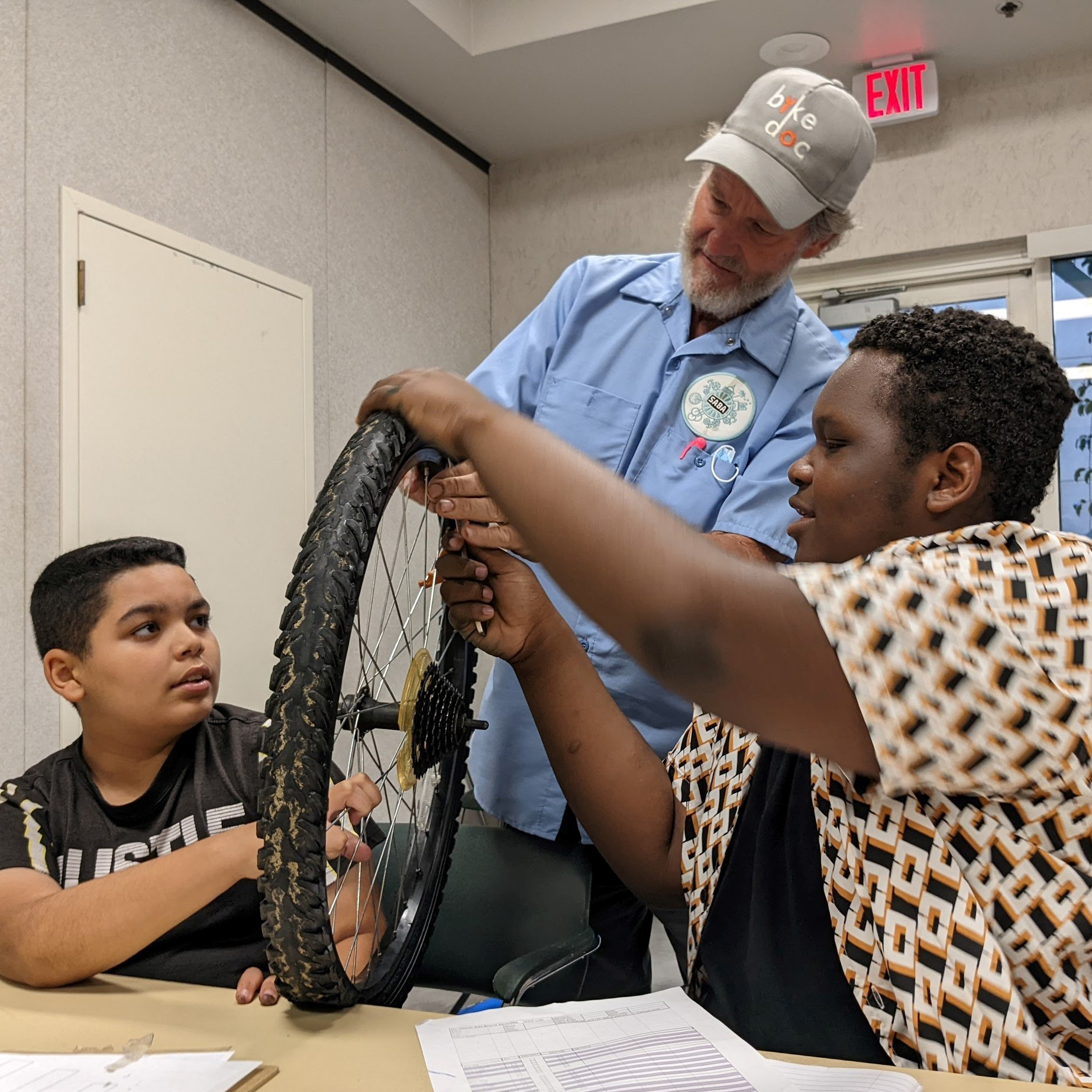 An older white man helps two young men of color fix a bike tire.