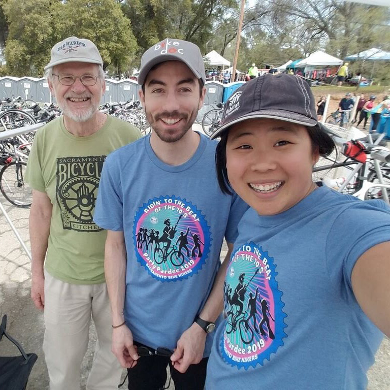 Three people - a young Asian woman, a young white man, and an older white man with bikes parked behind them.