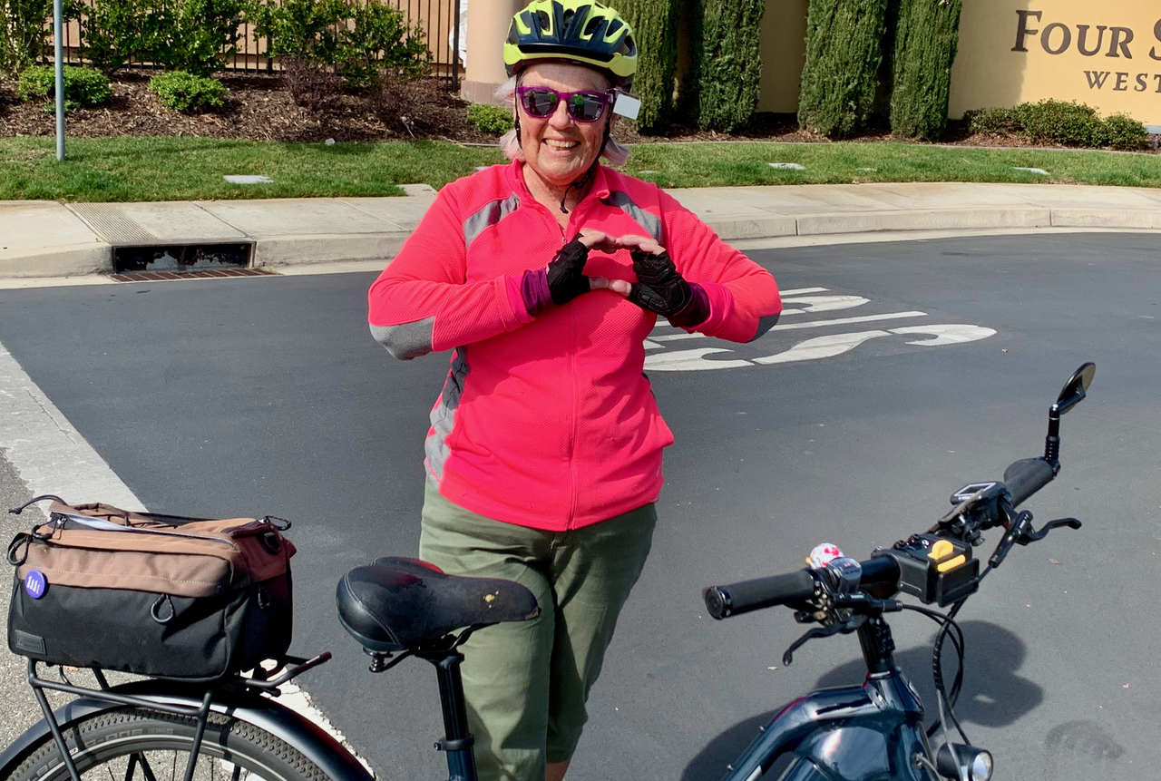 A woman wearing a yellow bike helmet and a pink jacket stands by a bike.