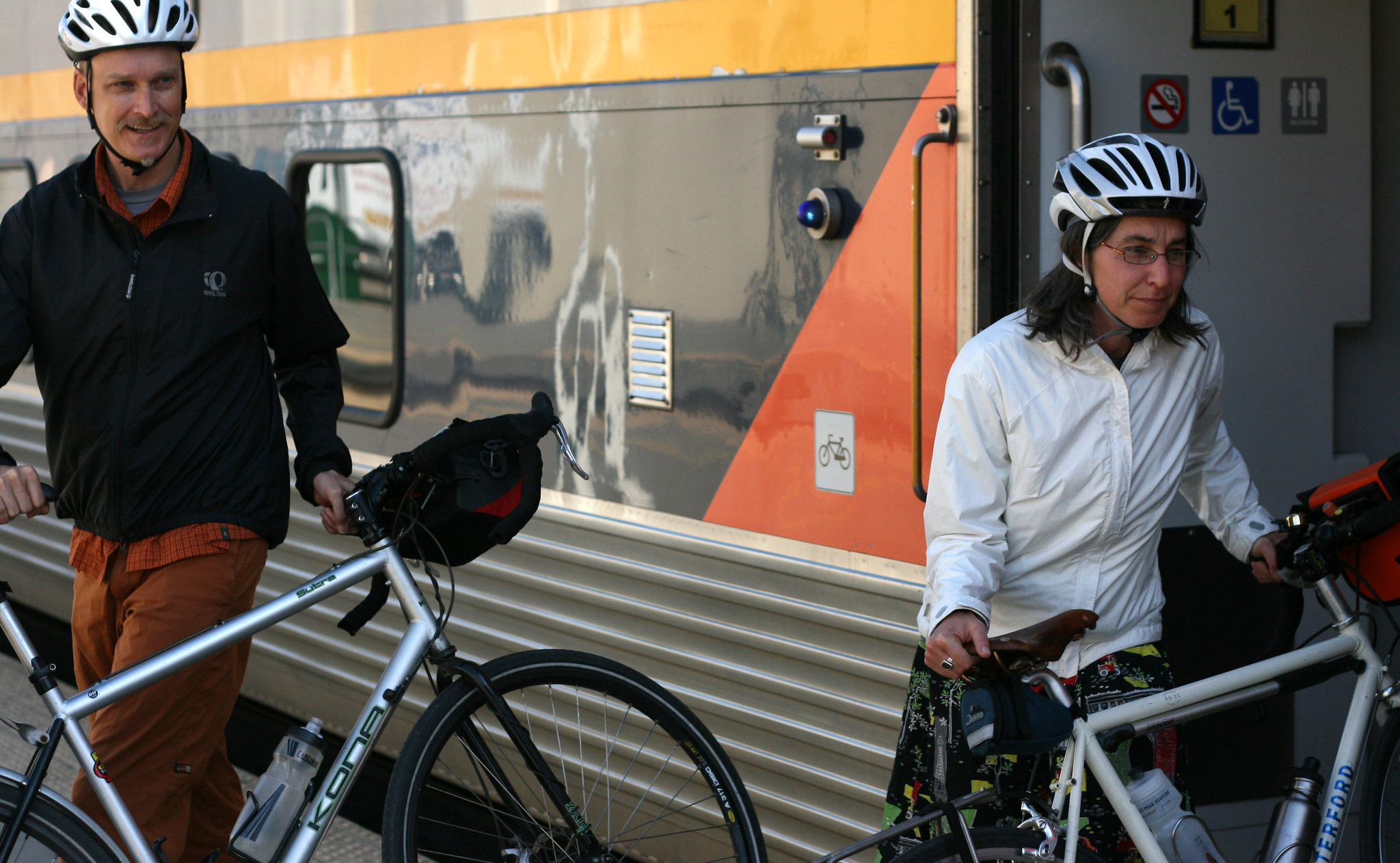 A man and a woman cyclist hold their bikes next to an Amtrak train.