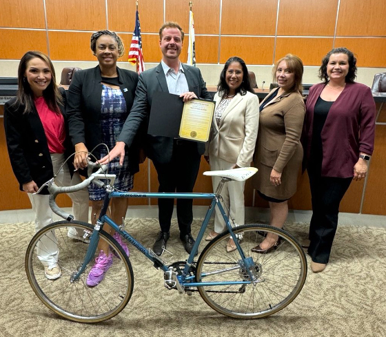 Six people pose in city council chambers, one man is holding a bike in front of him.