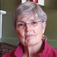 White woman with short gray hair wearing a red top and glasses.