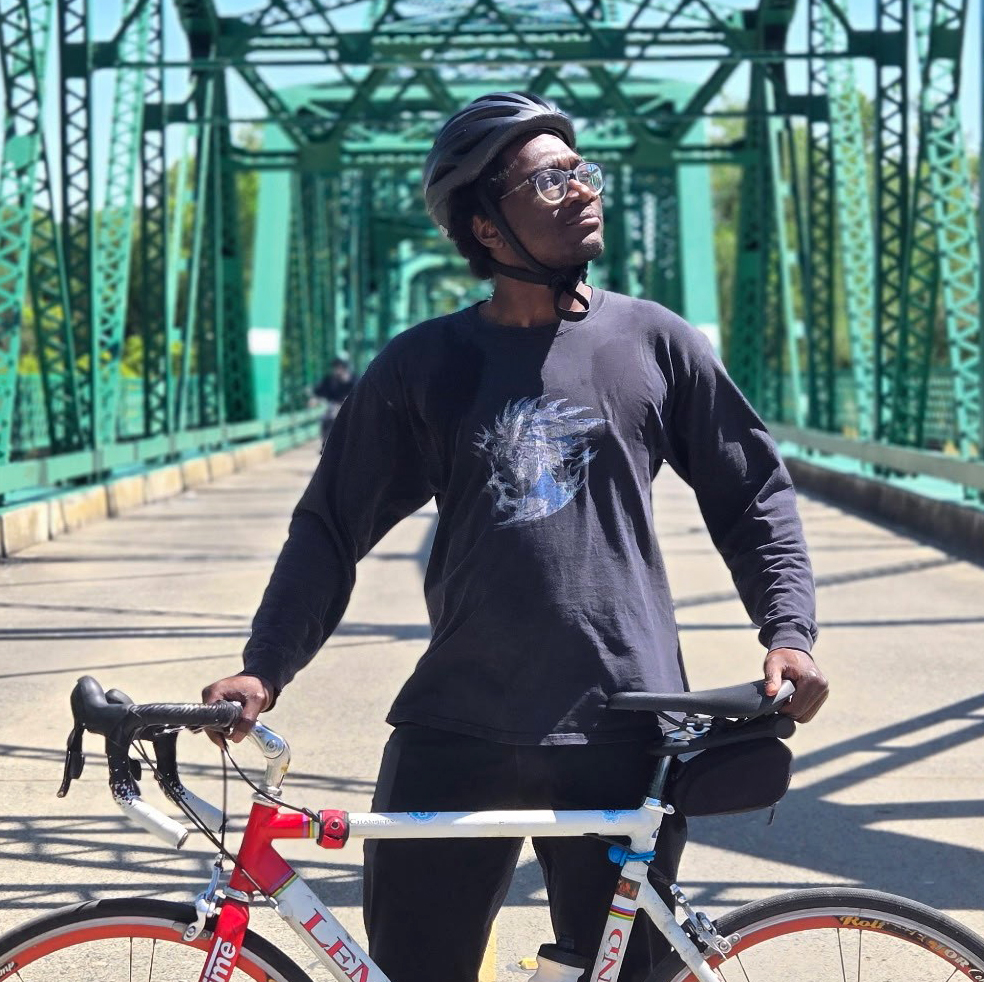 A man wearing a bike helmet stands next to his bike on a metal bridge.