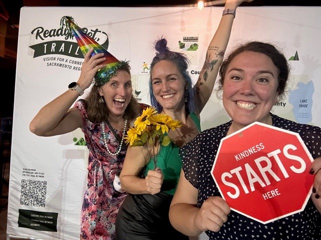 Three women, one wearing a party hat laugh as they ham it up at Velo Ball. One holds a sign saying, "Kindness Starts Here."